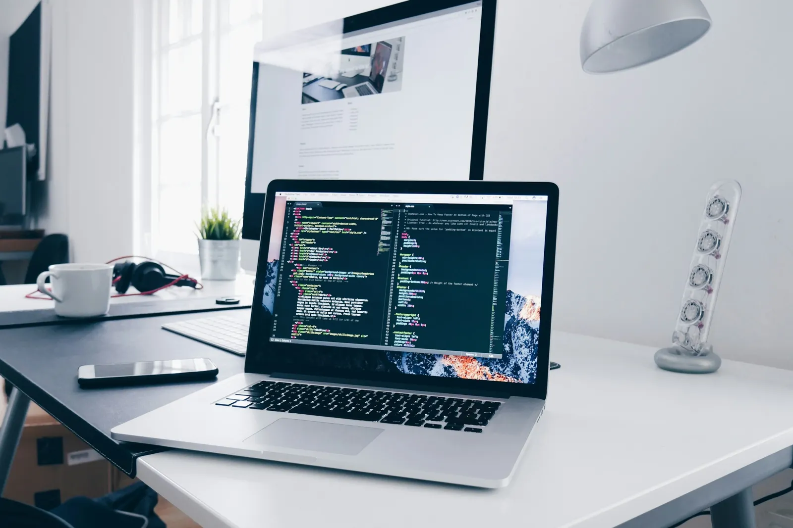 A laptop showing code on a tidy desk beside a large monitor.
